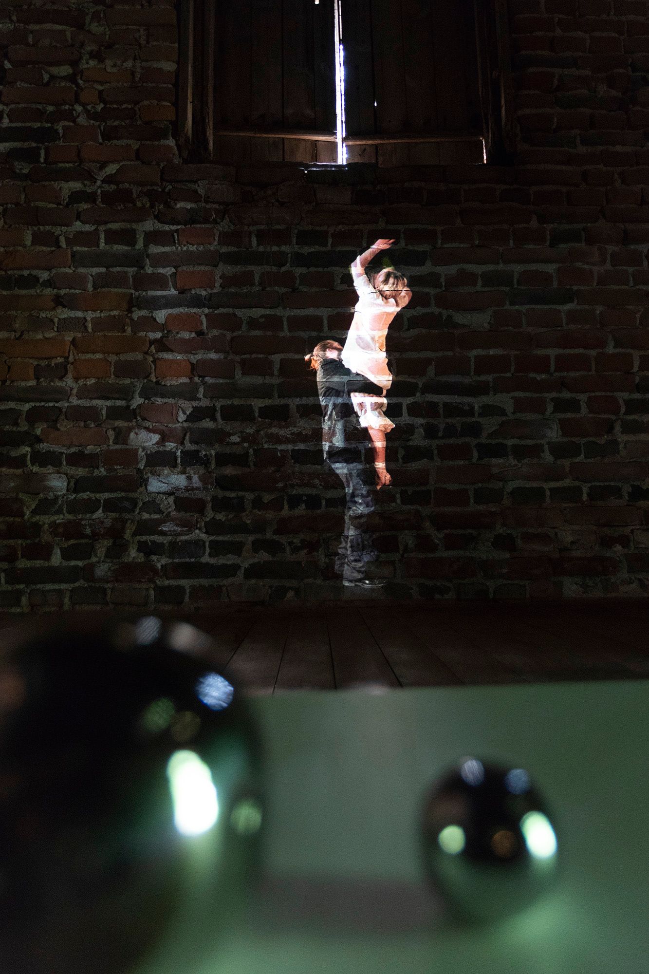 A picture with Mammu and Pasi being projected onto a brick wall, trying to reach a window above them. There are some shiny out-of-focus metallic balls in the foreground.
