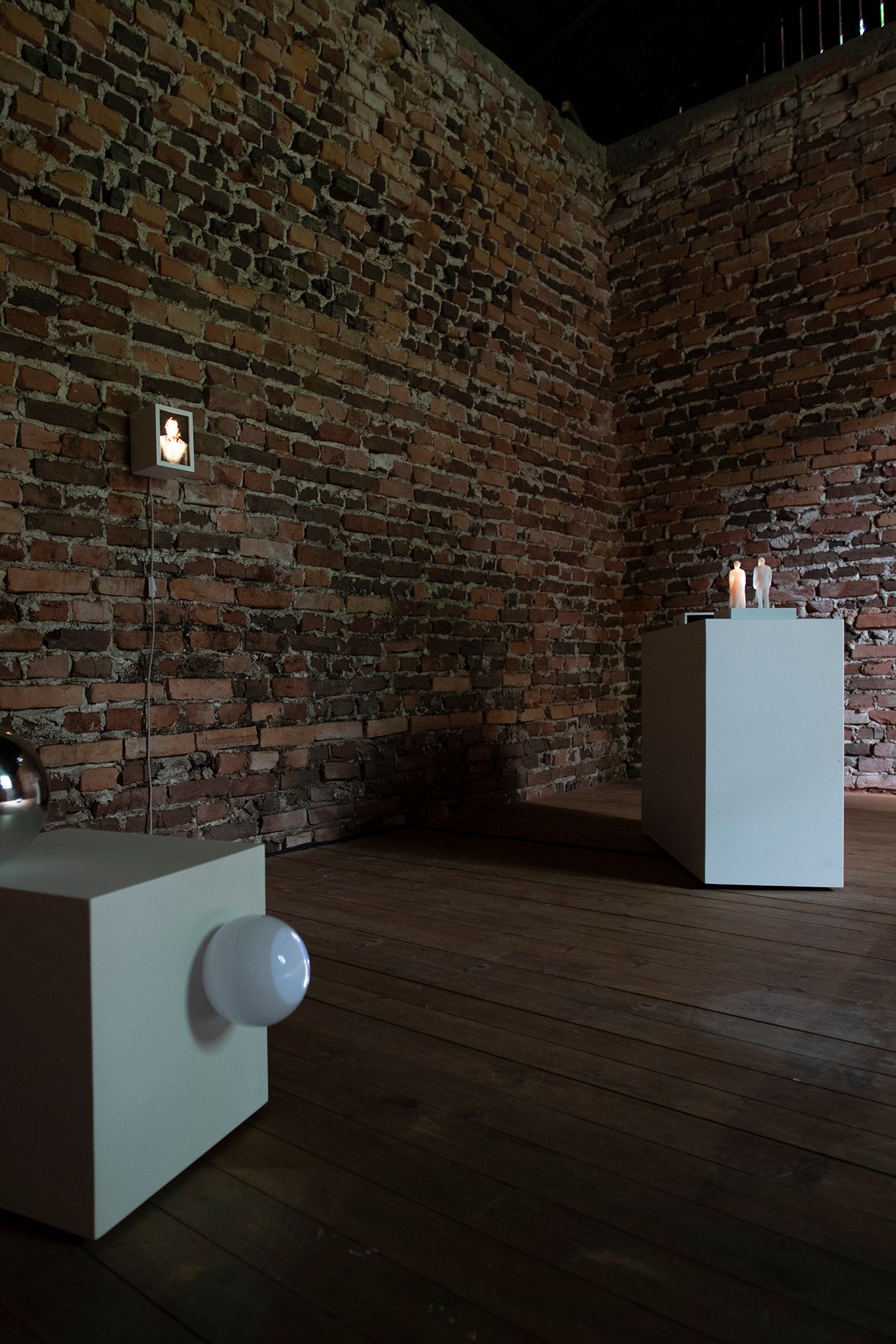 A general picture from the exhibition containing two white sculpture stands, the other one with some balls attached to it with small projections of Mammu and Pasi on them, the other one with white figures of Mammu and Pasi standing with their own pictures being projected onto them. There's a lightbox on the wall that has a picture of Mammu and Pasi overlaid on top of each other.