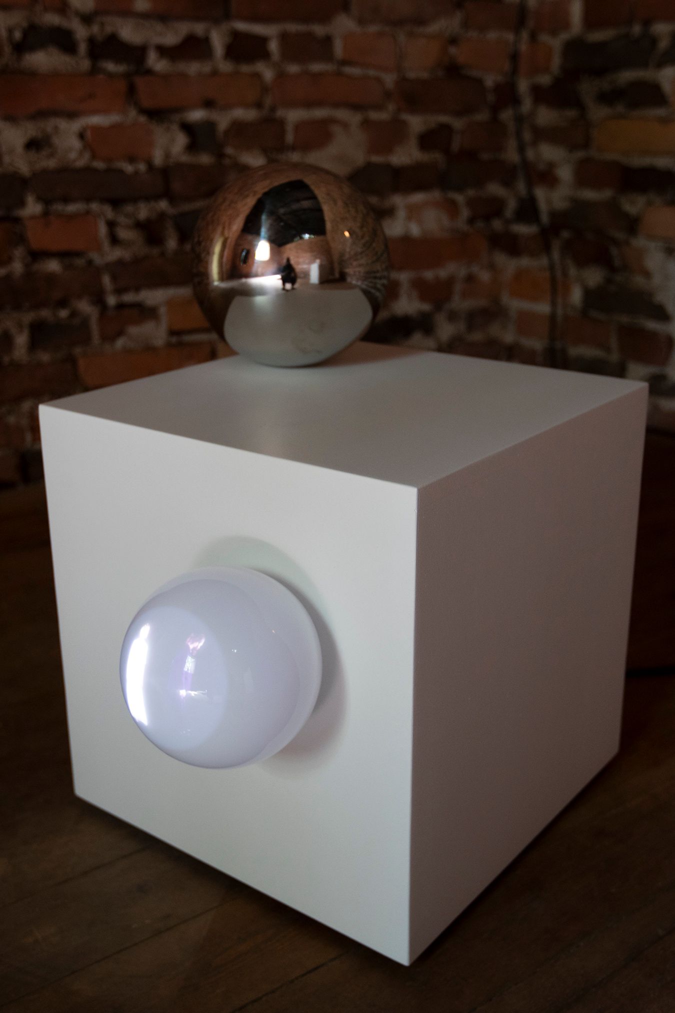A close-up shot of the white cube with a metallic ball on top of it and a frosted glass one attached to the side, with Mammu and Pasi standing being projected inside it.