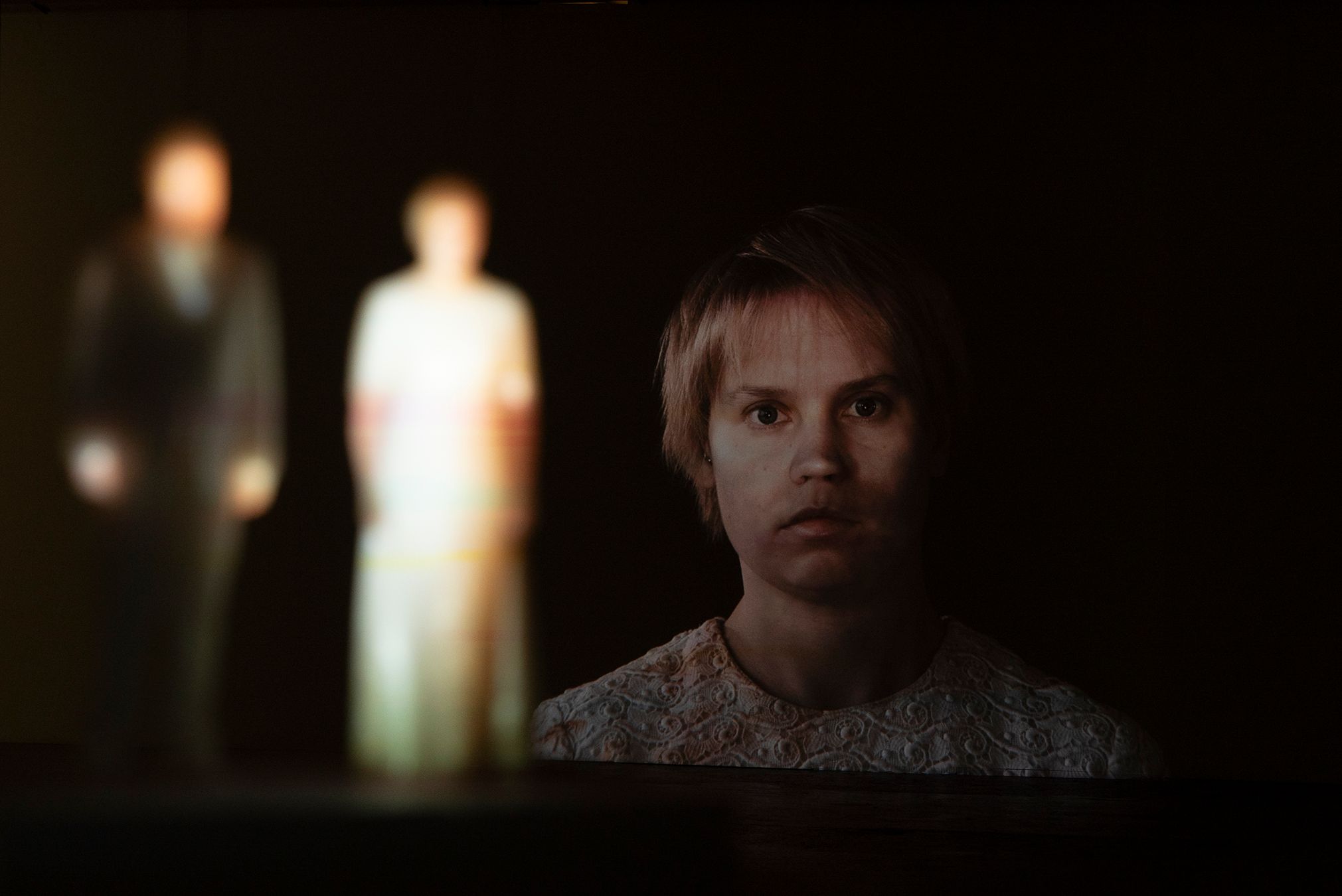 A picture with a projection of Mammu on the wall in the background, and small white figures of Mammu and Pasi with their pictures projected on them blurry in the foreground.