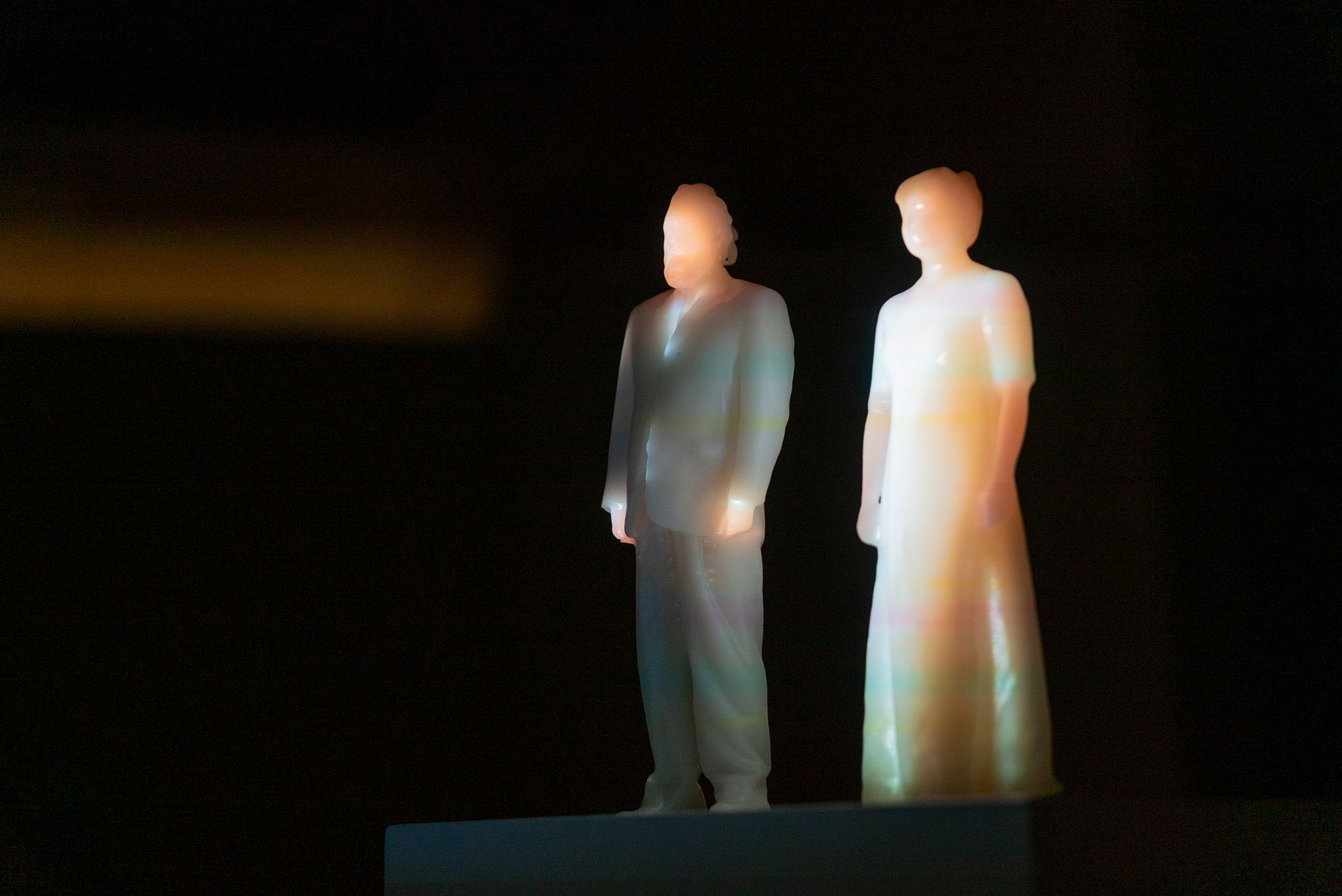 Plastic figures of Mammu and Pasi standing in a dark room, with a full-body projection of Mammu and Pasi on them.