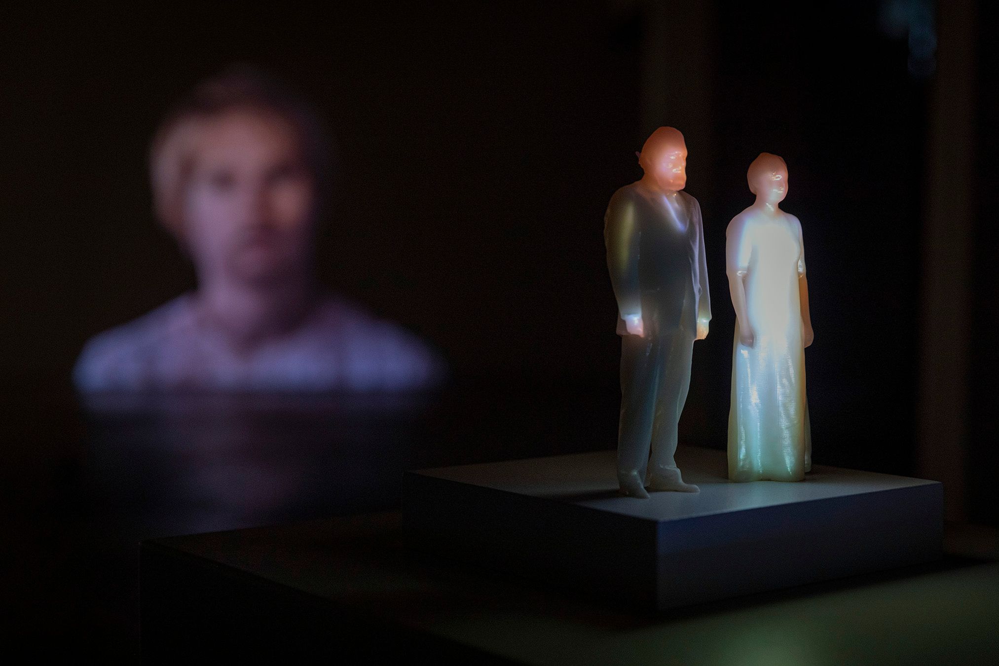 Plastic figures of Mammu and Pasi standing in a dark room, with a full-body projection of Mammu and Pasi on them.