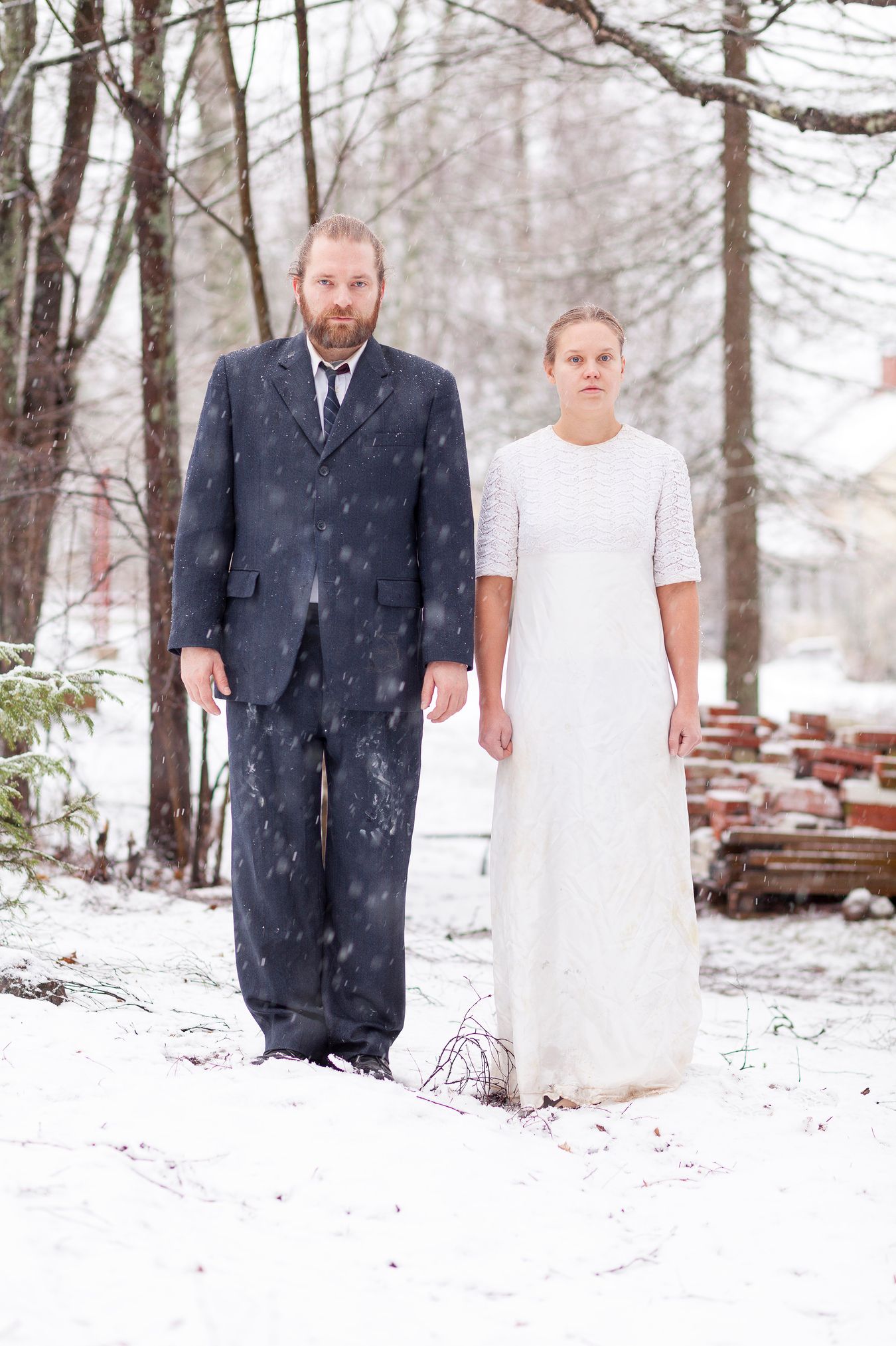 Mammu and Pasi standing in their wedding attire on their lawn with neutral expressions staring at the camera. It is winter and snow is falling. Their clothes are still relatively clean, with no stains of paint.