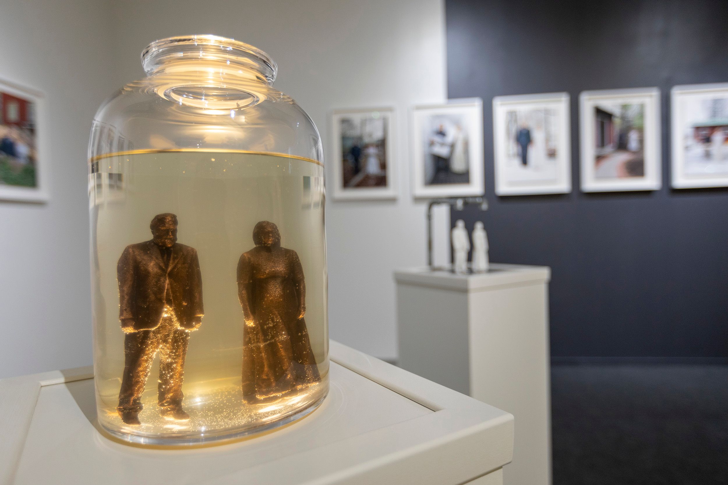A close-up picture of bronze figures of Mammu and Pasi standing inside a glass container filled with yellowish liquid, a young batch of kombucha.
