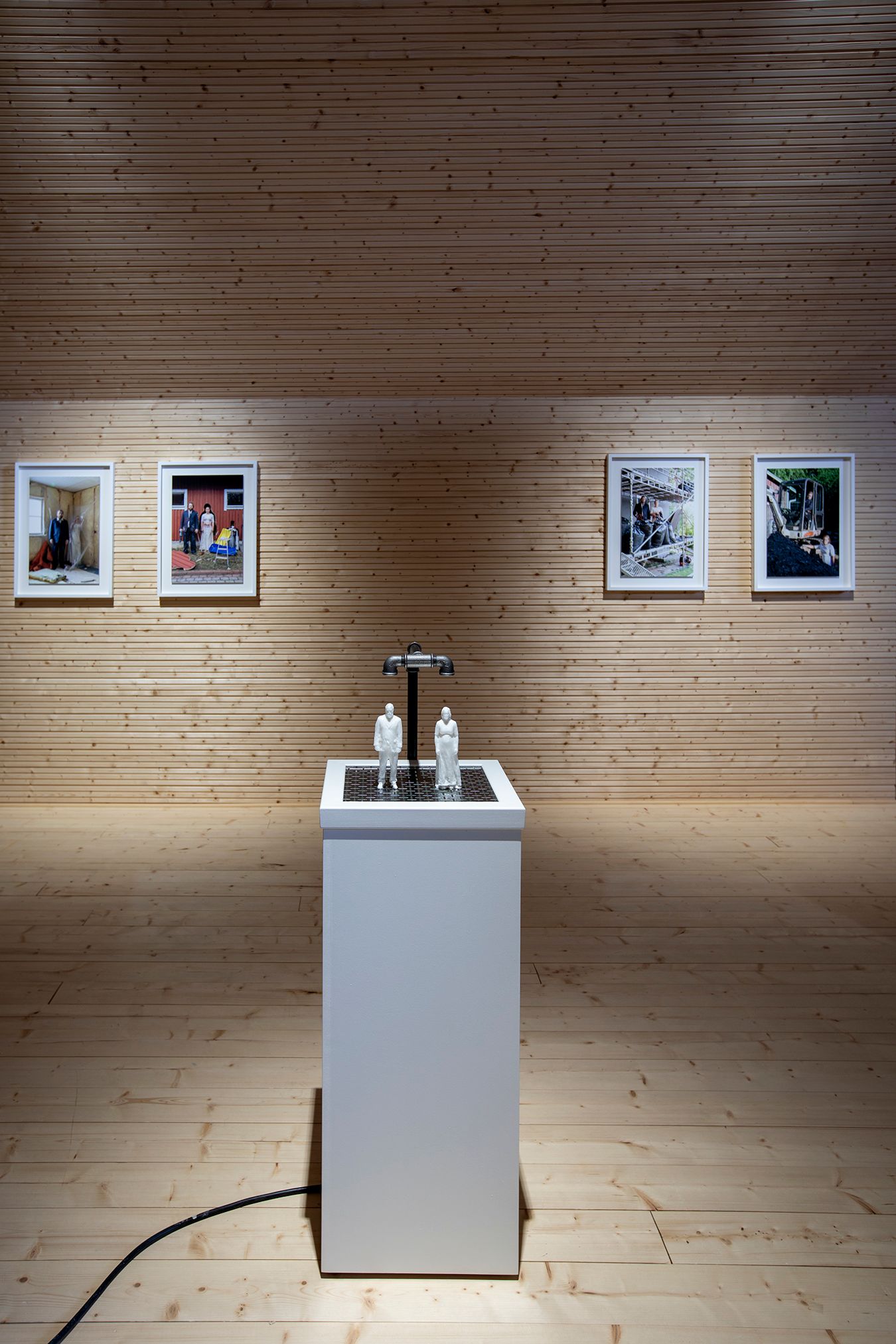 Picture of a white sculpture stand with soap figures of Mammu and Pasi under water faucets. In the background, some of the photographs in the series on a wooden wall.