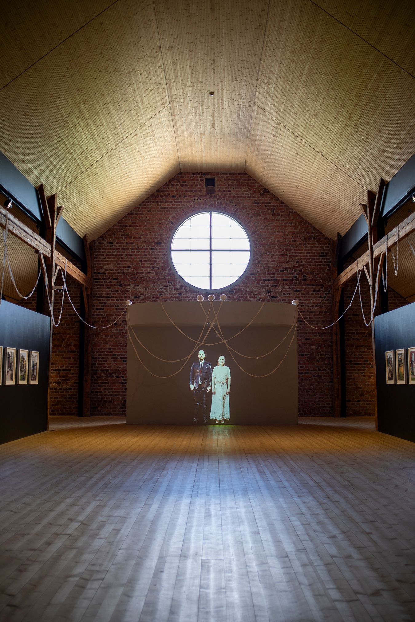 Overview picture from the exhibition; a well-lit room with projections of Mammu and Pasi, some photographs, and rag rug strips hanging from structural elements.