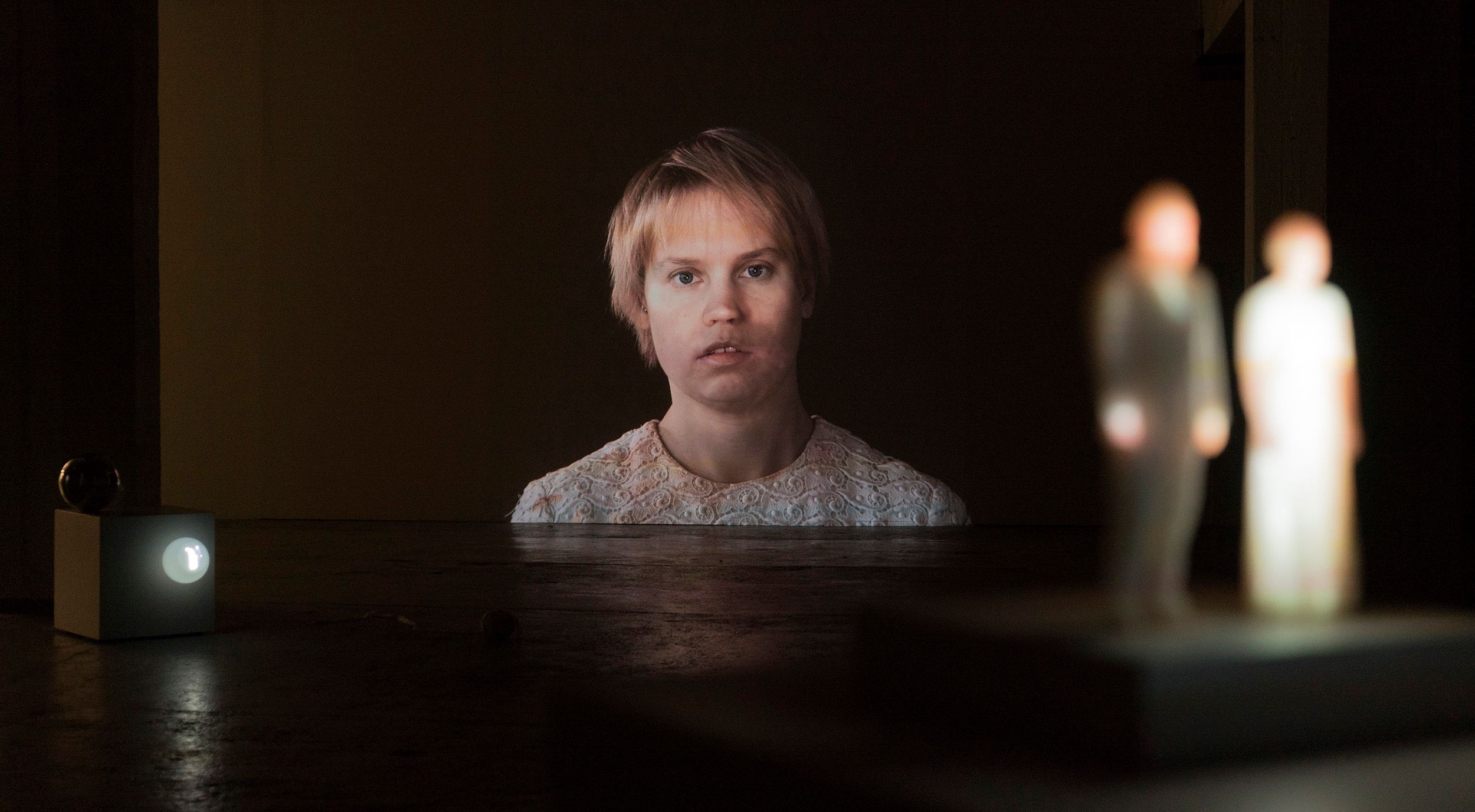Wide-angle view from an exhibition in a dark room with artworks from the project. Main subject is a projected video of Mammu with Pasi's face being projected on it.