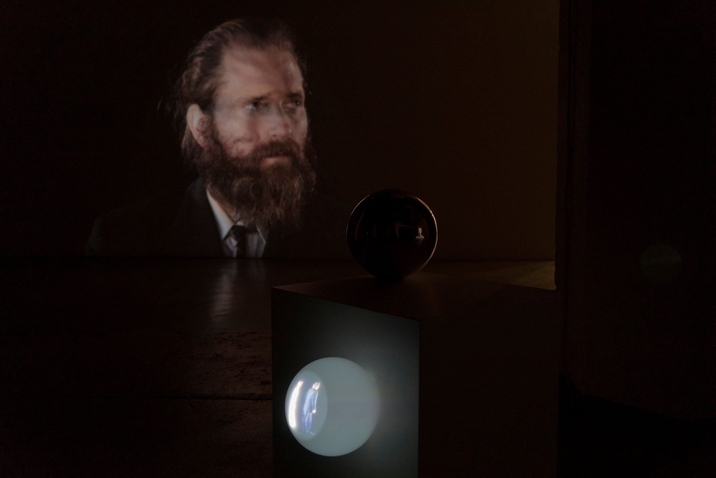 Close-up view from an exhibition in a dark room with artworks from the project. Main subject is a cube with two spheres hanging on it, one of which has a video projection of Pasi and Mammu.