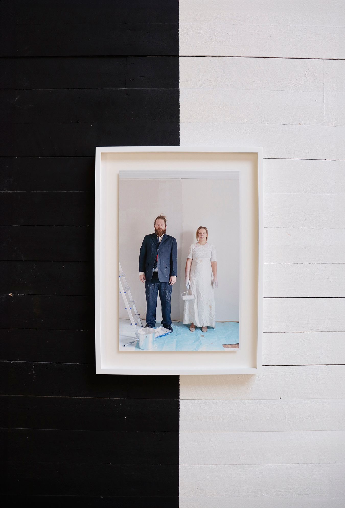 Close-up picture of a photograph of Mammu and Pasi hanging on a wooden wall. The wall is painted black and white, with the photograph having been hung just at the edge of those two areas. The edge of black and white is repeated in the photograph, where Mammu and Pasi are standing in front of a wall that is in process of being painted, creating a visible edge of paint between them.