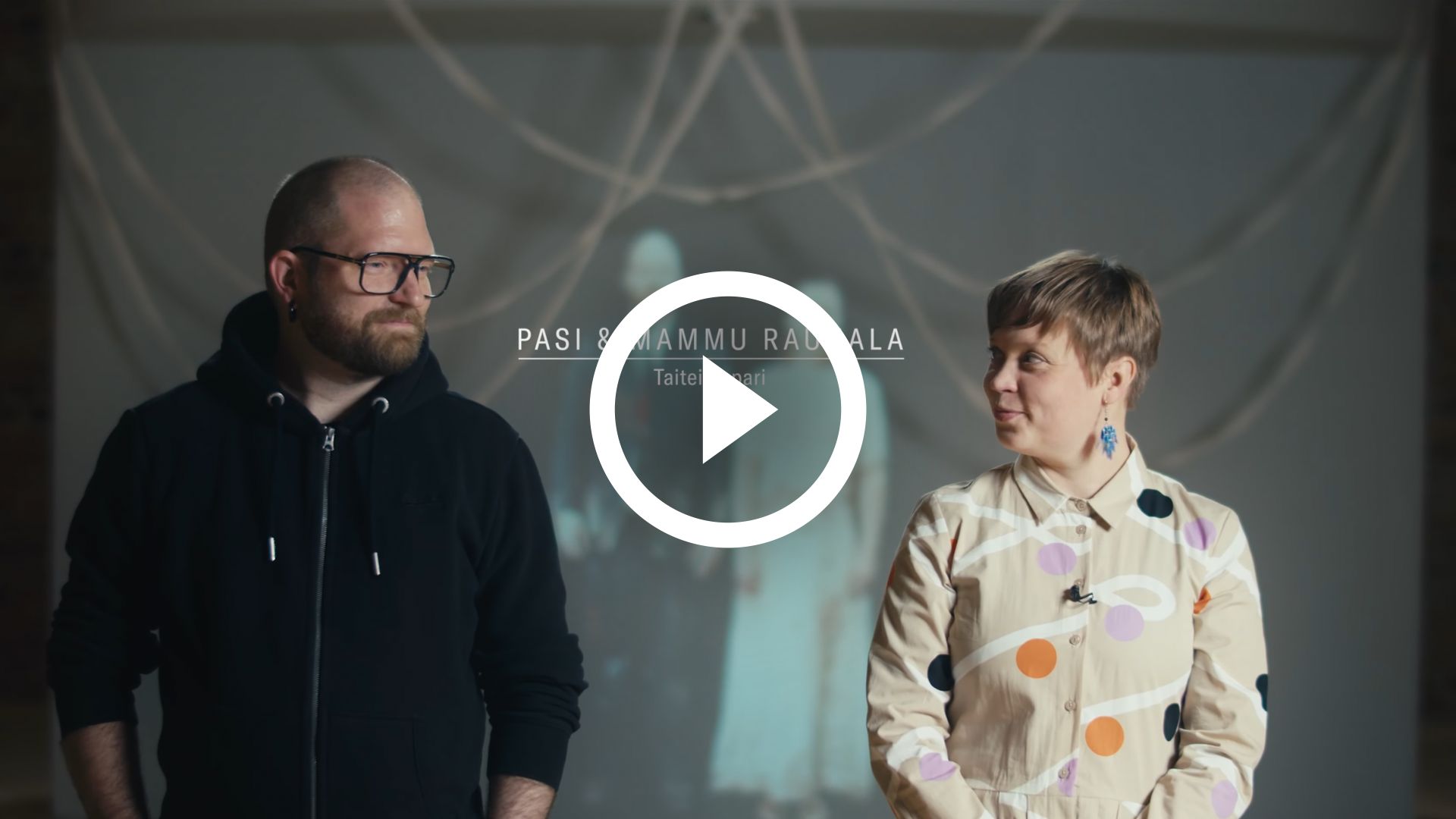 Play video. Picture of mammu and Pasi standing together in an interview.