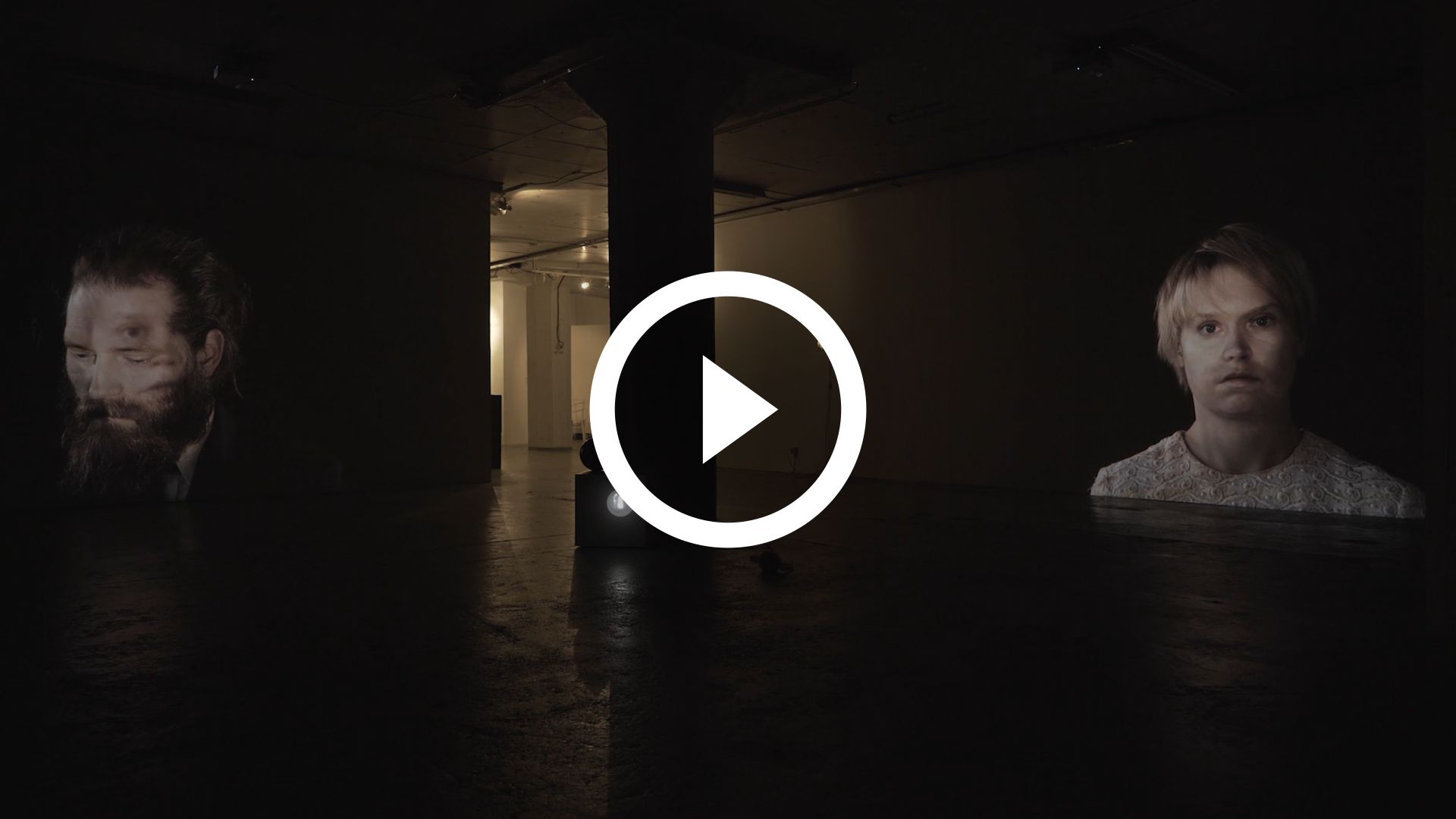 Play video. Picture of dark exhibition room with projections of Mammu and Pasi with each other projected on their faces.